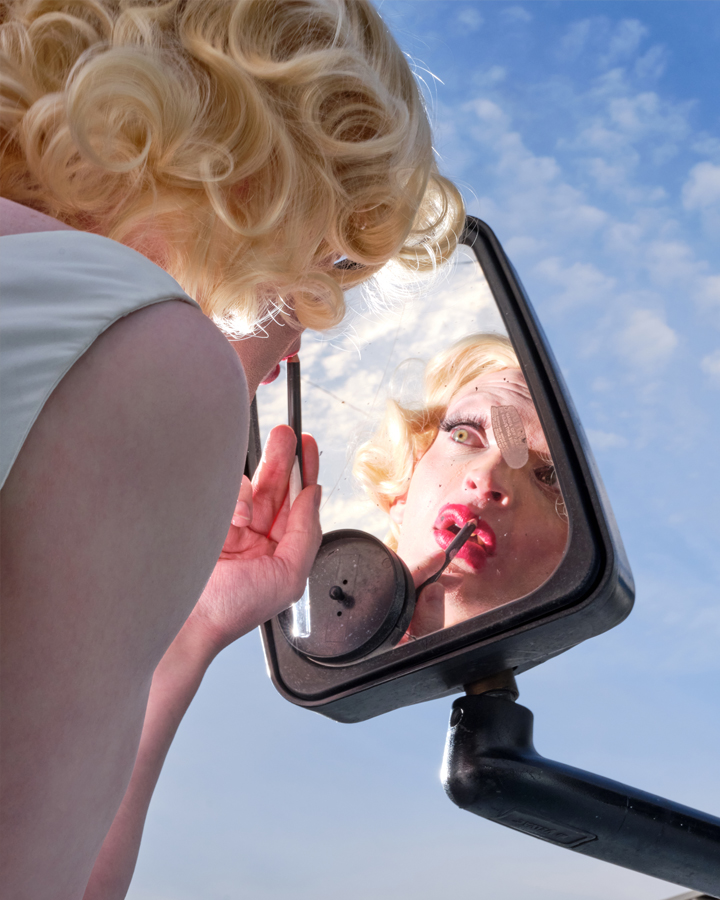 Brooks Ginnan dressed as marilyn monore fixing her lipstick in the rear view mirror of a van with a blue sky in the background